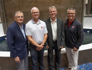 A group of four adults standing in front of a water feature with a tiled wall. The individuals are dressed in business or semi-formal attire, with some wearing suits and others in blazers and UW Badgers polo shirts.