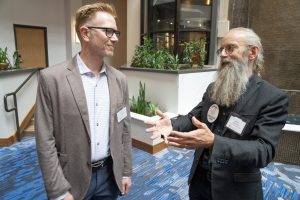 Two adult men stand together talking and one is gesturing.