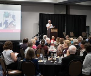 A adult person stands in front of a room full of people sitting at tables. He is holding a microphone and talking with a screen about his family behind him.