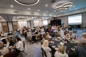 A full hotel banquet room with tables of people sitting together and a stage and podium at the front of the room.