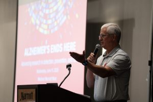 A man stands at a podium holding a microphone and gesturing. A screen behind him reads alzheimers ends here.