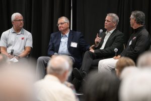 Four adults sit in chairs on a stage and one holds a microphone.